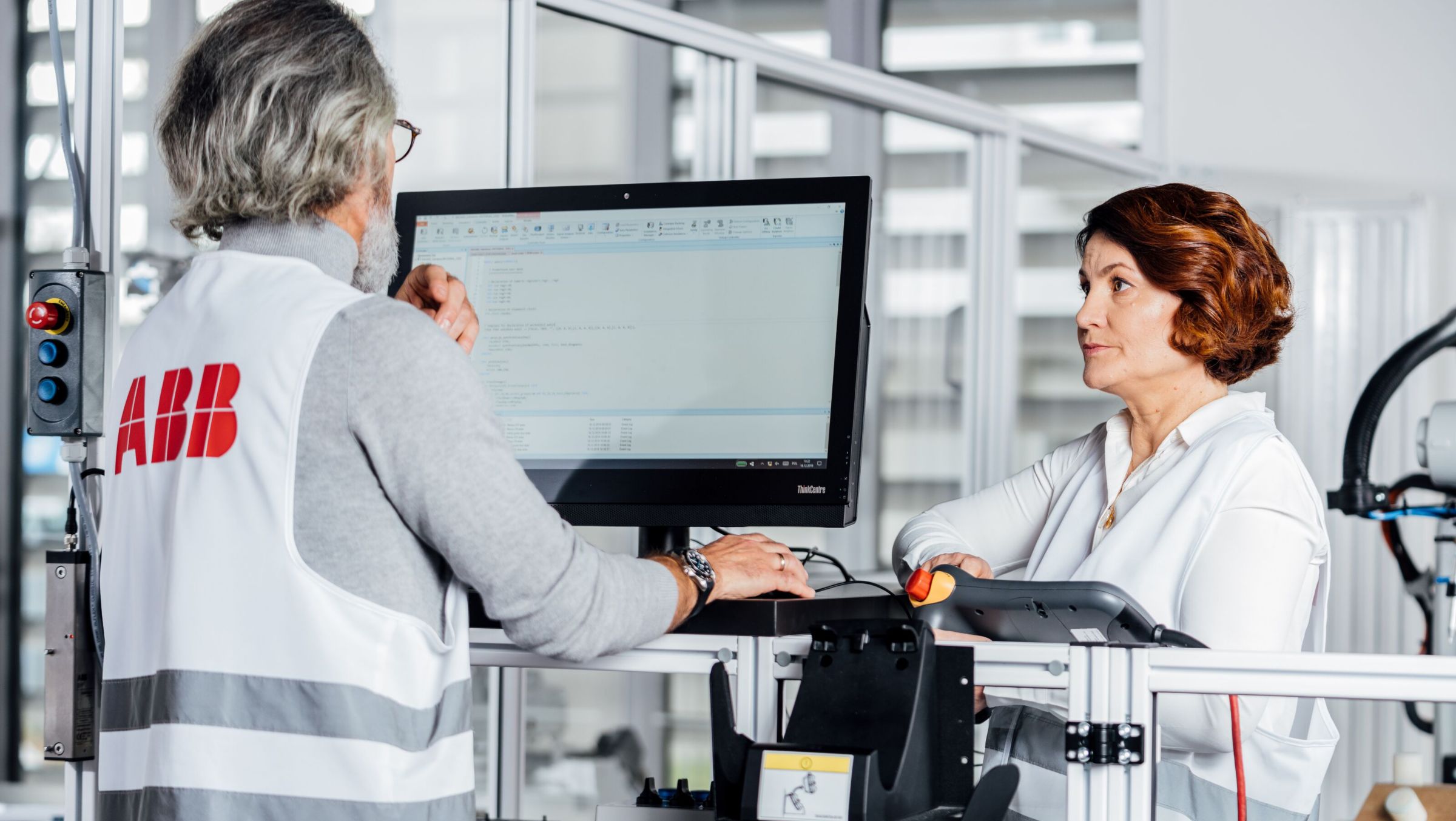 Two individuals collaborate at a workstation, analyzing data on a computer in a modern industrial setting. The environment is bright and organized.
