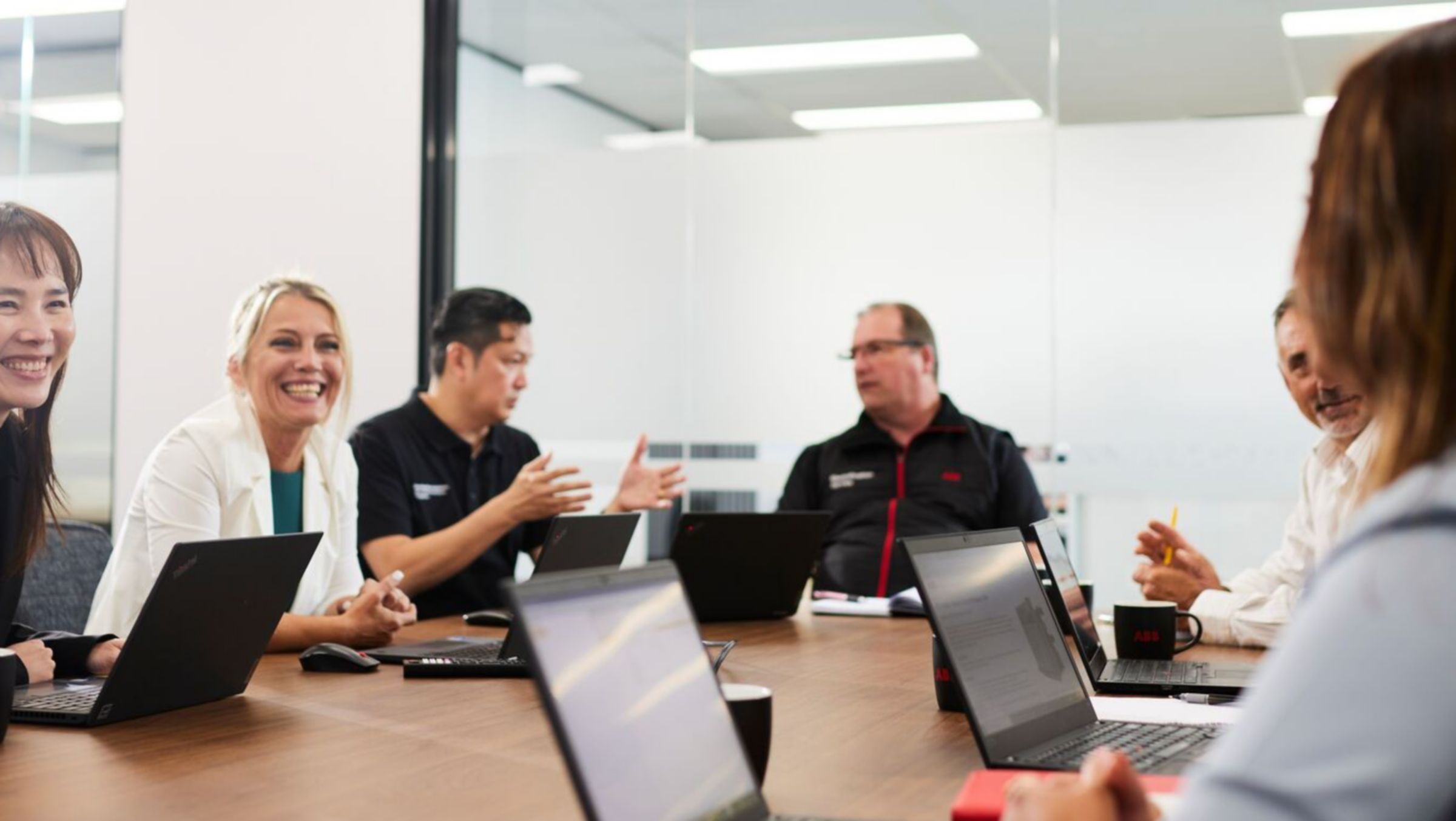 A group of colleagues sit around a long wooden table in a glass-walled meeting room, engaged in a collaborative discussion with open laptops and notebooks spread before them.