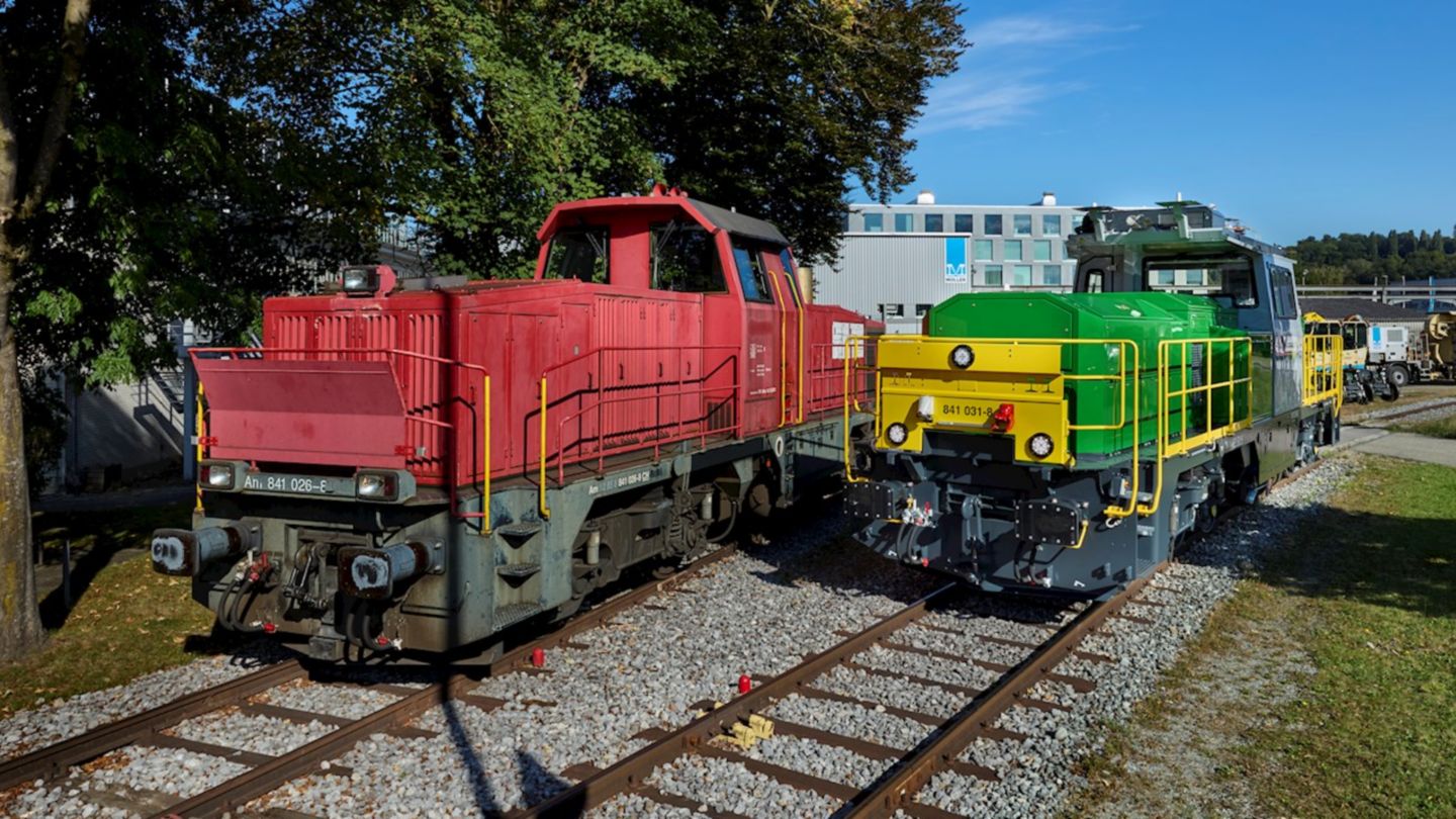 Dos locomotoras diésel, una roja y otra verde, colocadas sobre vías con árboles y un edificio al fondo bajo un cielo azul claro.