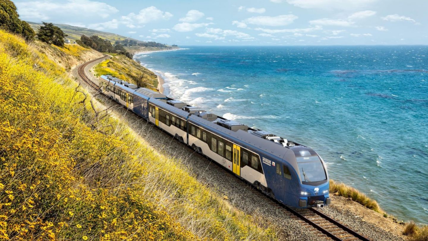 Un tren azul y blanco viaja a lo largo de pintorescas vías costeras, flanqueadas por vibrantes flores amarillas y el brillante océano azul.
