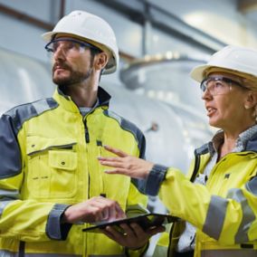 Three Heavy Industry Engineers Stand in Pipe Manufacturing Factory, Use Digital Tablet Computer, Have Discussion. Large Pipe Assembled. Design and Construction of Oil, Gas and Fuels Transport Pipeline