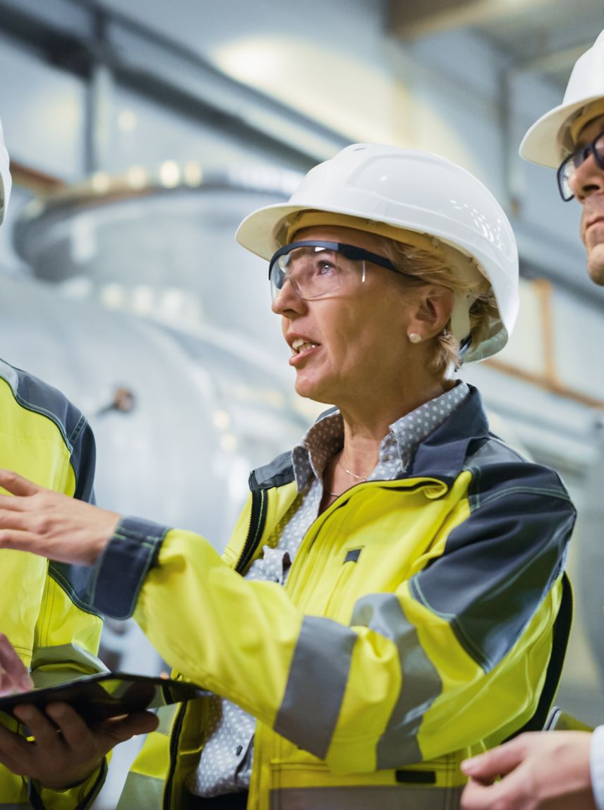 Three Heavy Industry Engineers Stand in Pipe Manufacturing Factory, Use Digital Tablet Computer, Have Discussion. Large Pipe Assembled. Design and Construction of Oil, Gas and Fuels Transport Pipeline